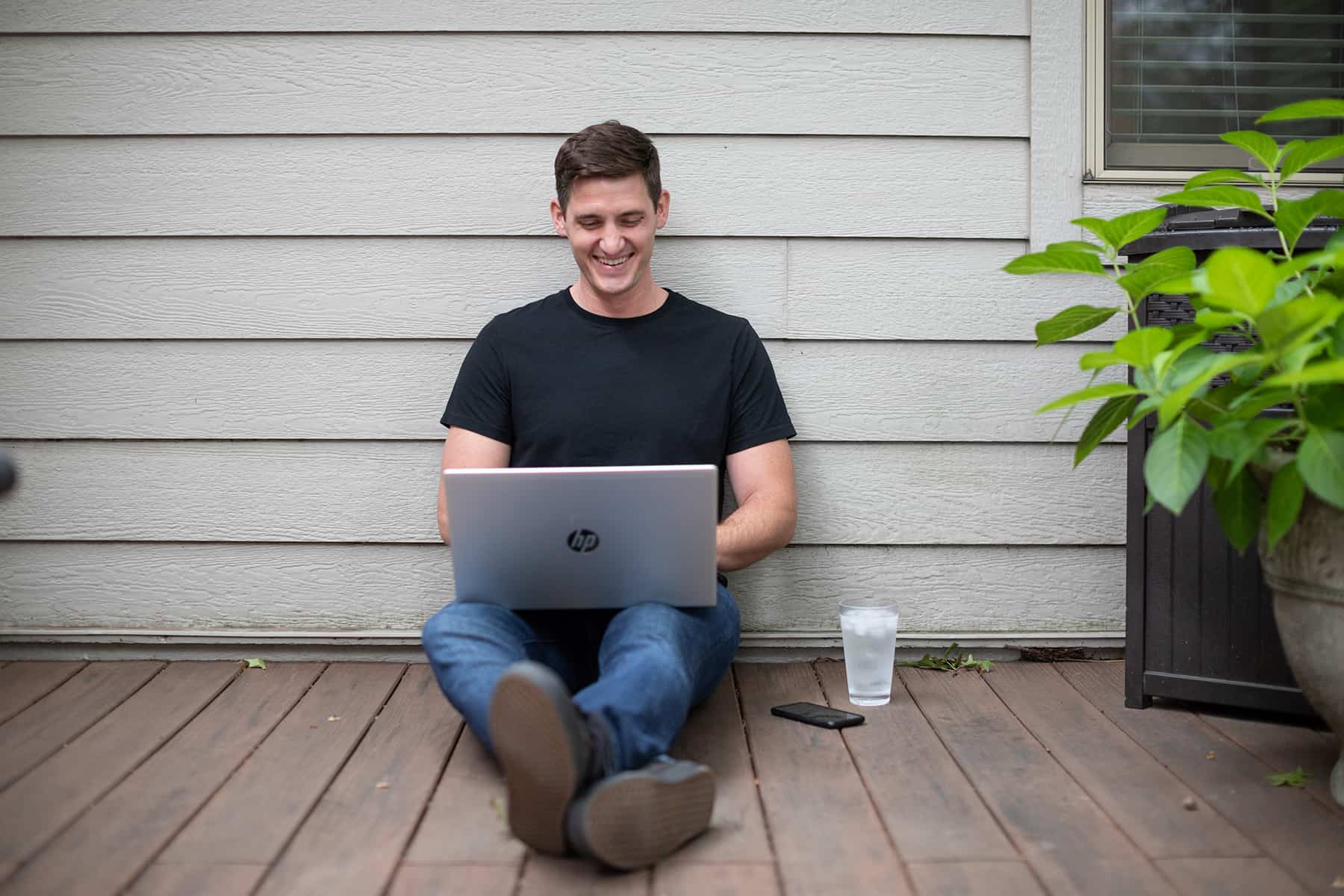 smiling young man sitting on a porch and working on his laptop and drinking a glass of water because he is thirsty for IT knowledge