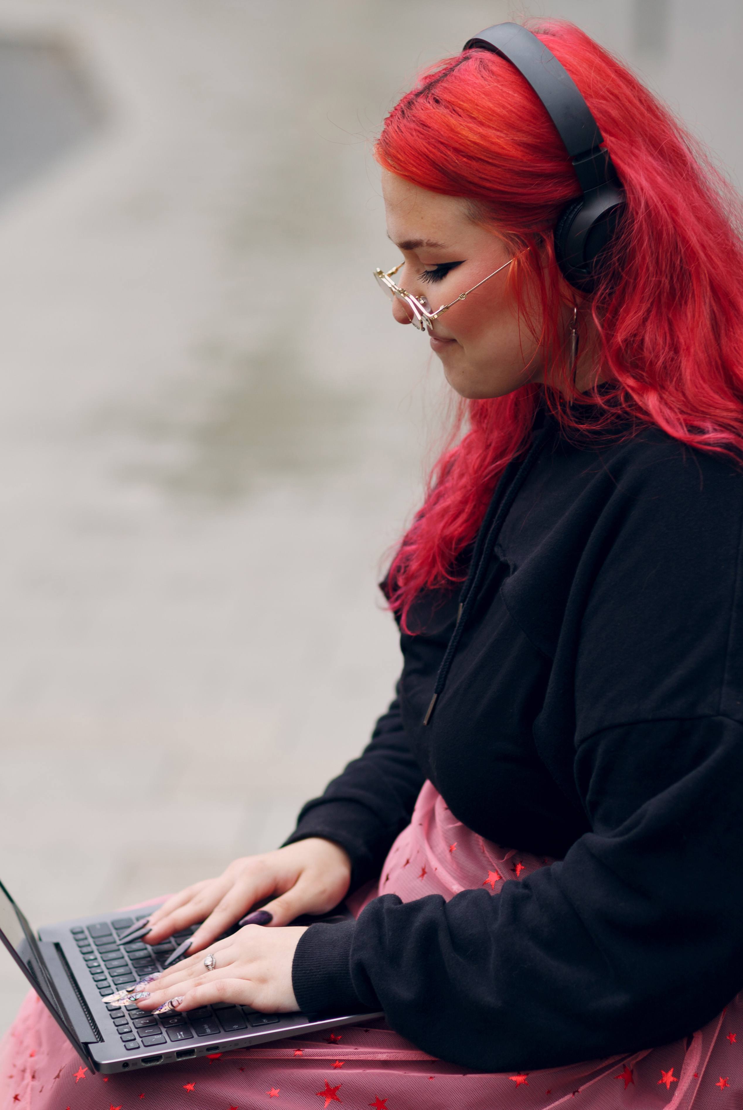 Caucasian plus-sized woman sits outdoors on a bench with a laptop on her lap. She has bright red, dyed hair, and wears glasses and headphones.