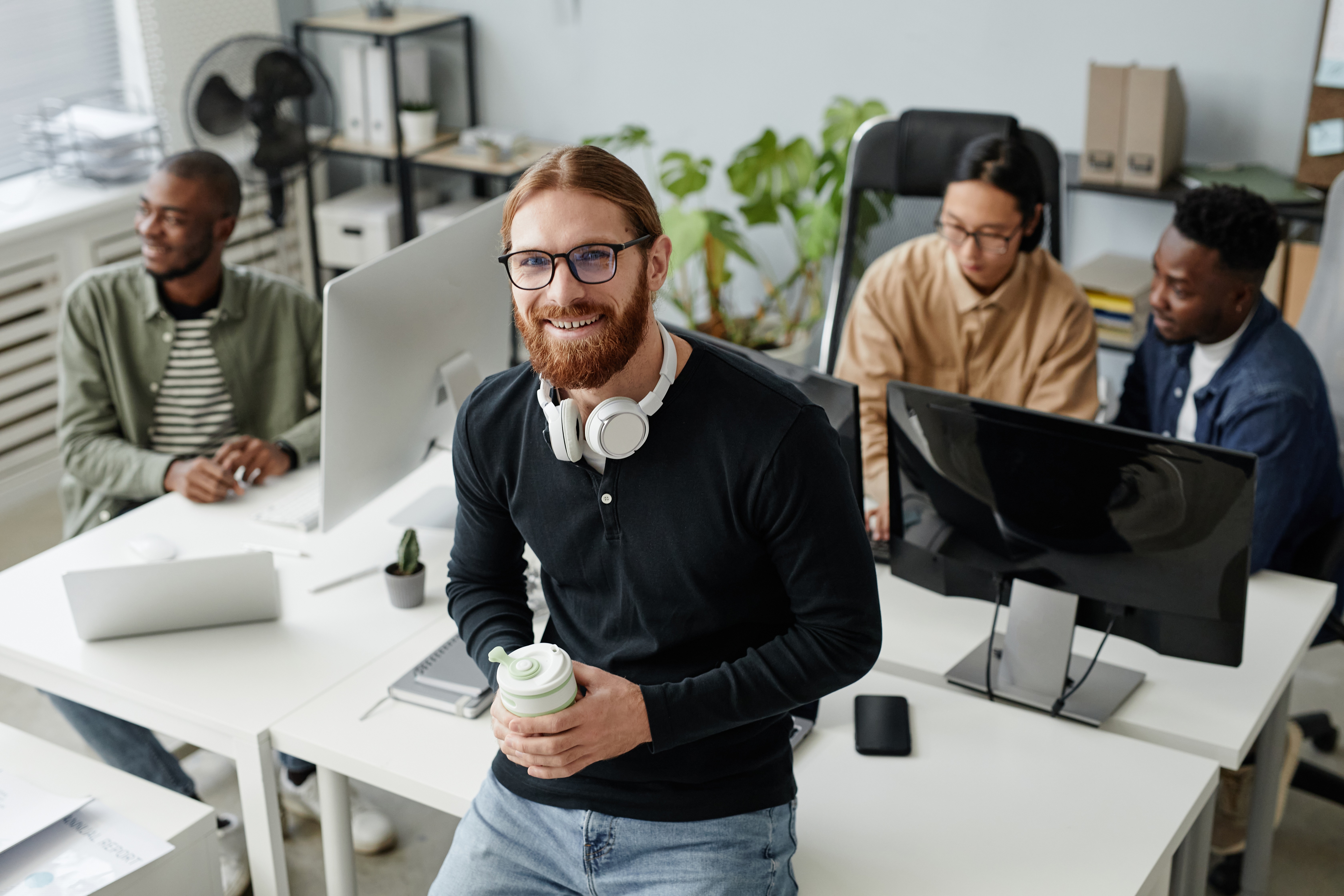 Smiling bearded man in casual attire wearing glasses and headphones stands with coffee in hand. Behind him is a coworking desk with three diverse men collaborating at desktop computers